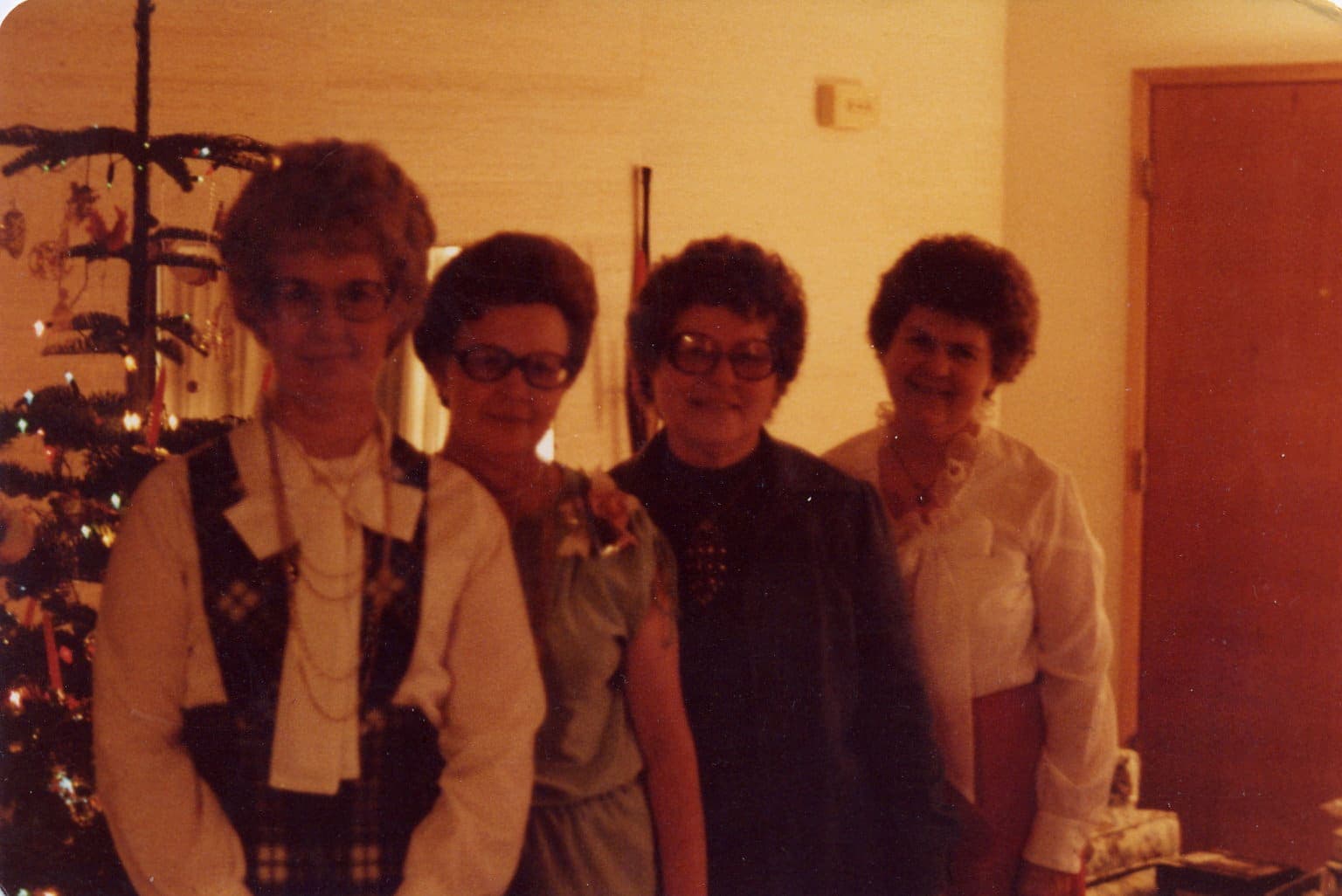 Four women standing together, dressed in vintage attire, in a warmly lit room with a Christmas tree in the background.
