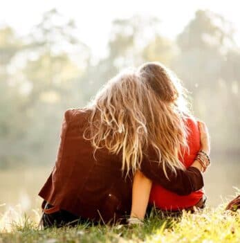 Two friends sit closely together by a calm waterway, enjoying each other's company.