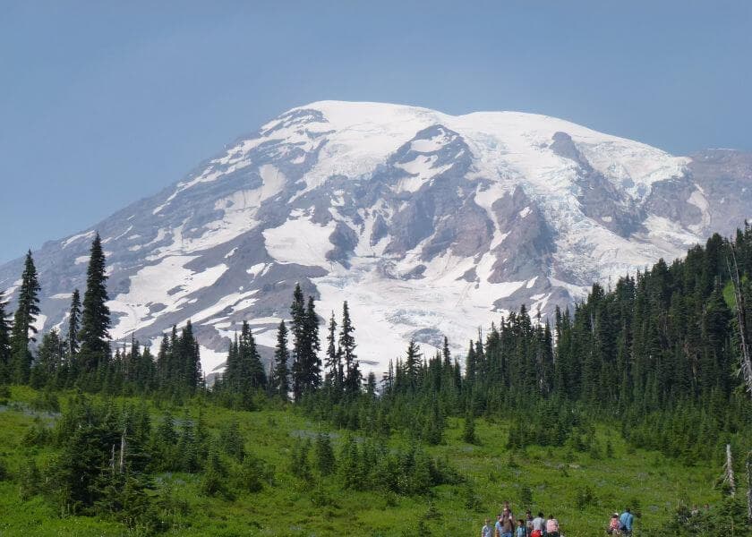 A snow-capped mountain rises above a forested landscape with hikers in the foreground.