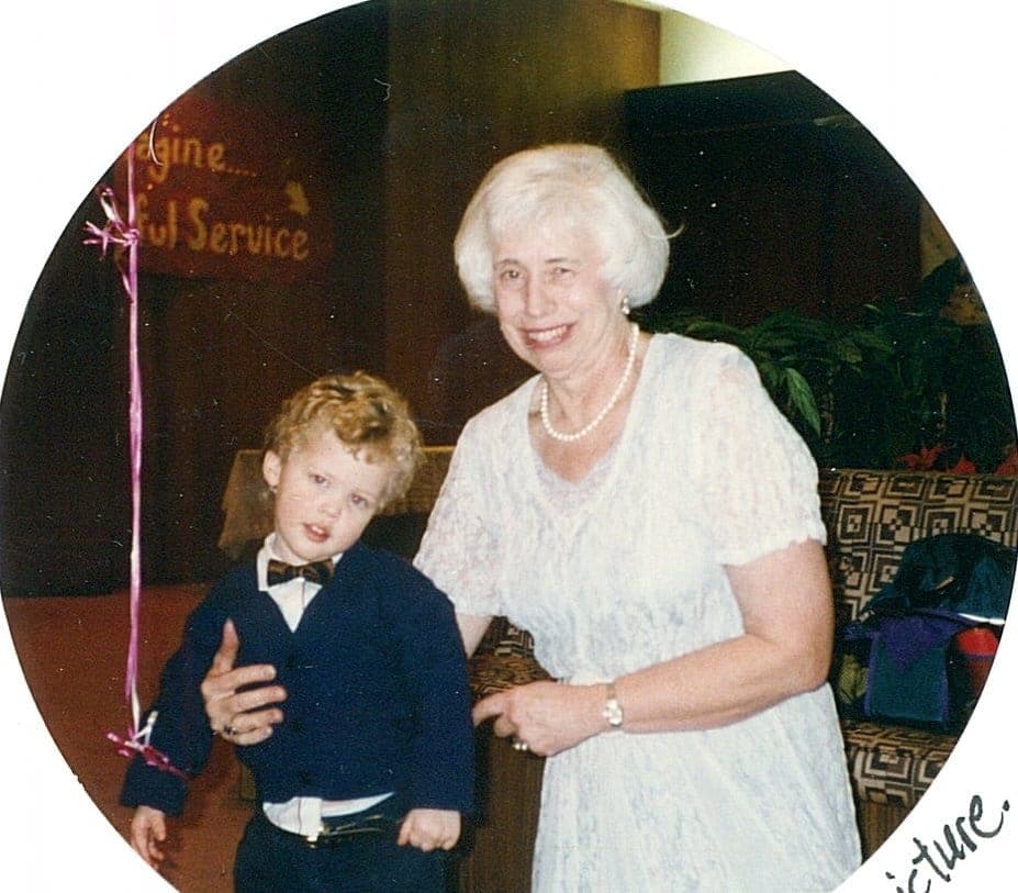 A smiling elderly woman stands next to a young boy in formal attire, both posing for a photo at a festive event.