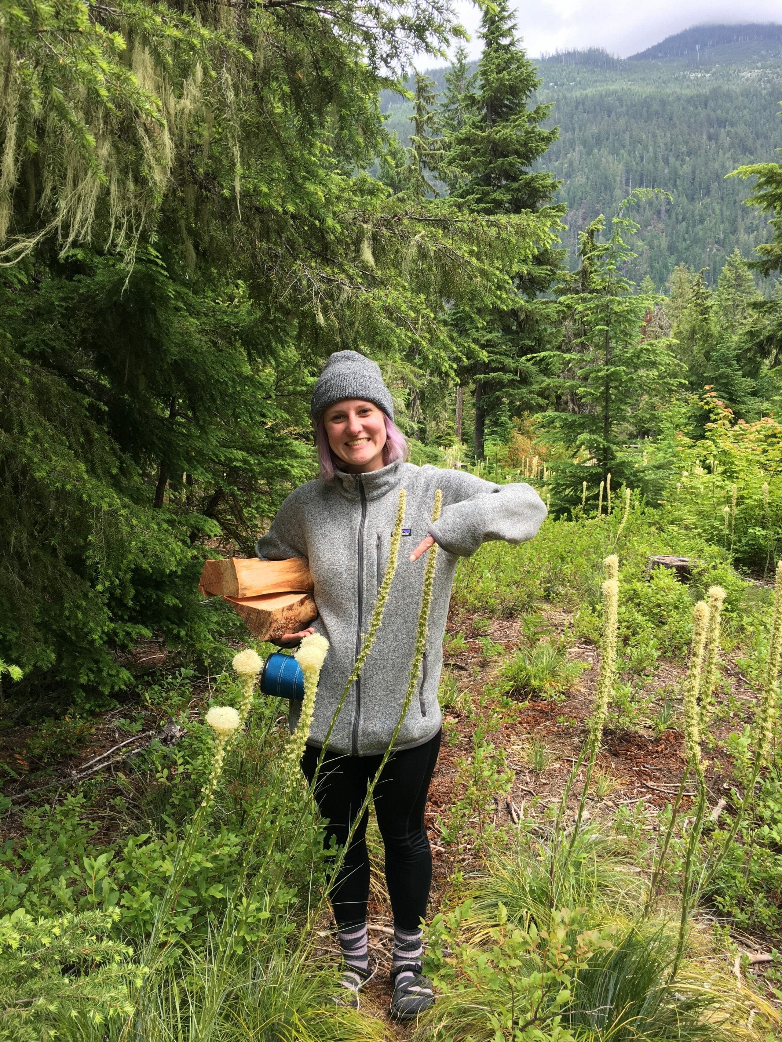 A smiling person in a gray sweater and hat stands in a lush green forest, holding firewood and pointing with one hand.