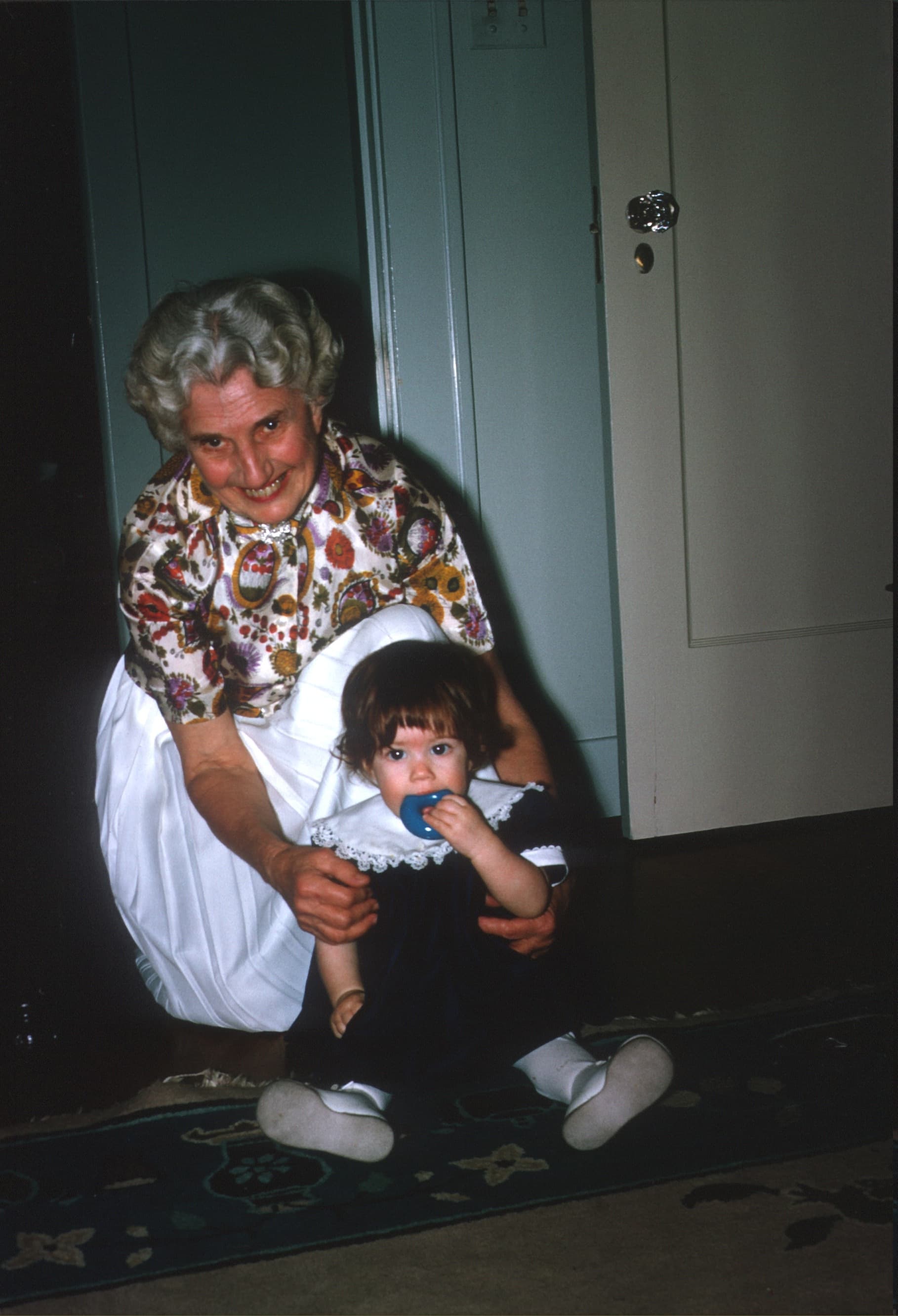 A smiling elderly woman crouches beside a young child sitting on the floor, both looking at the camera.
