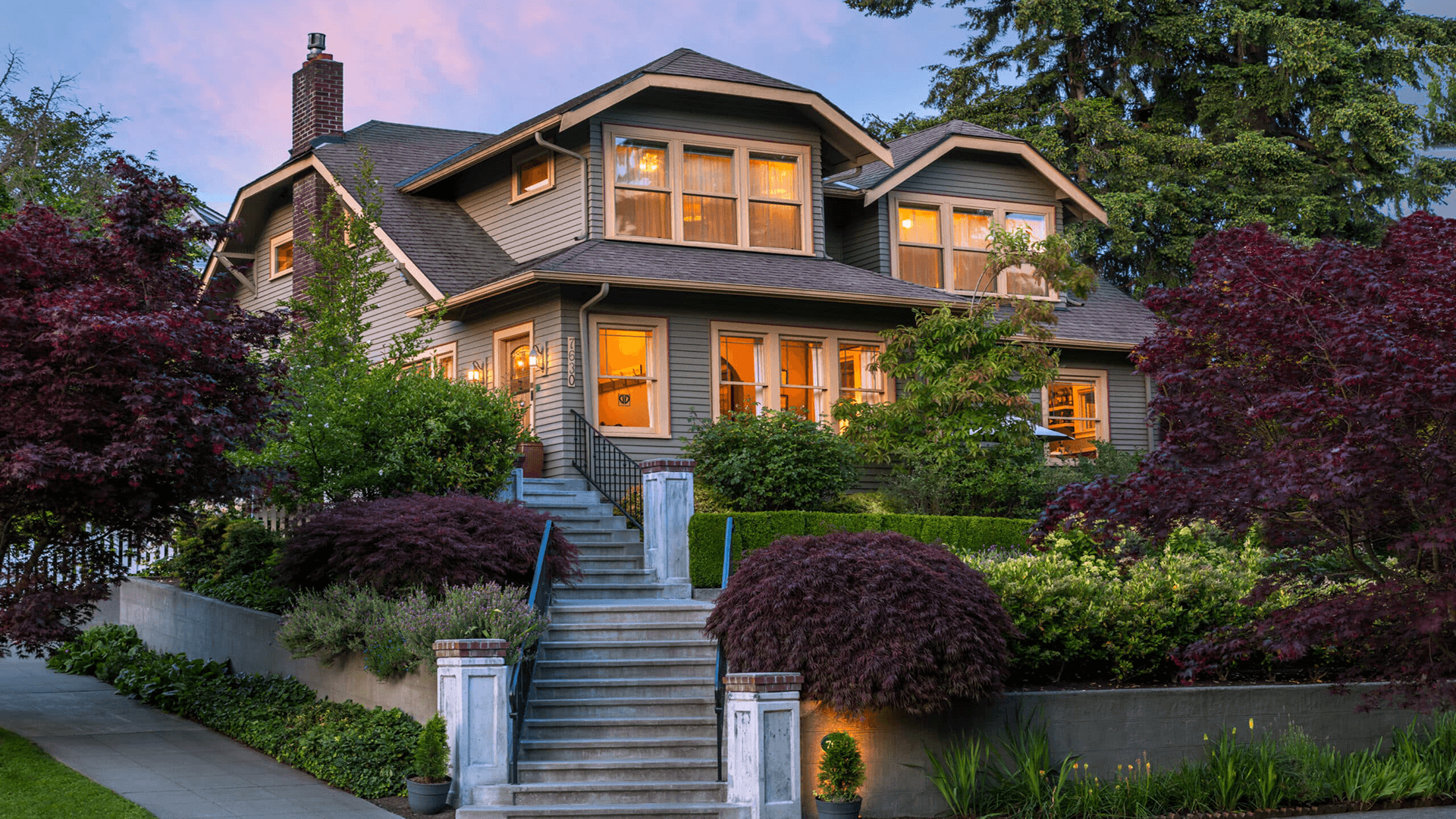 A charming two-story house surrounded by lush greenery and colorful bushes.