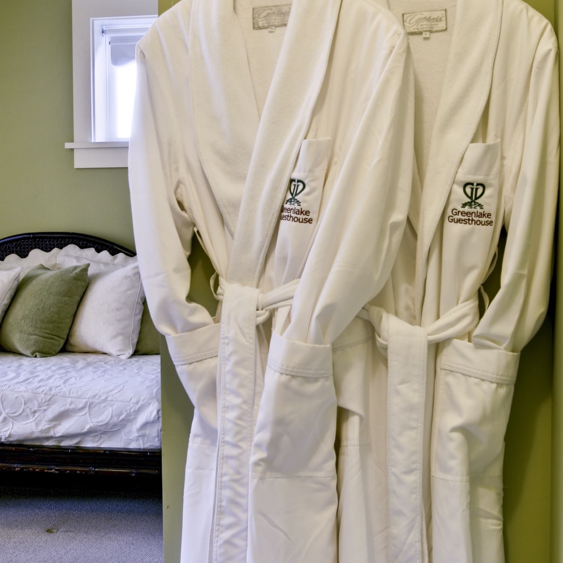 Two white bathrobes hang on a wall next to a bed with decorative pillows in a cozy guestroom.