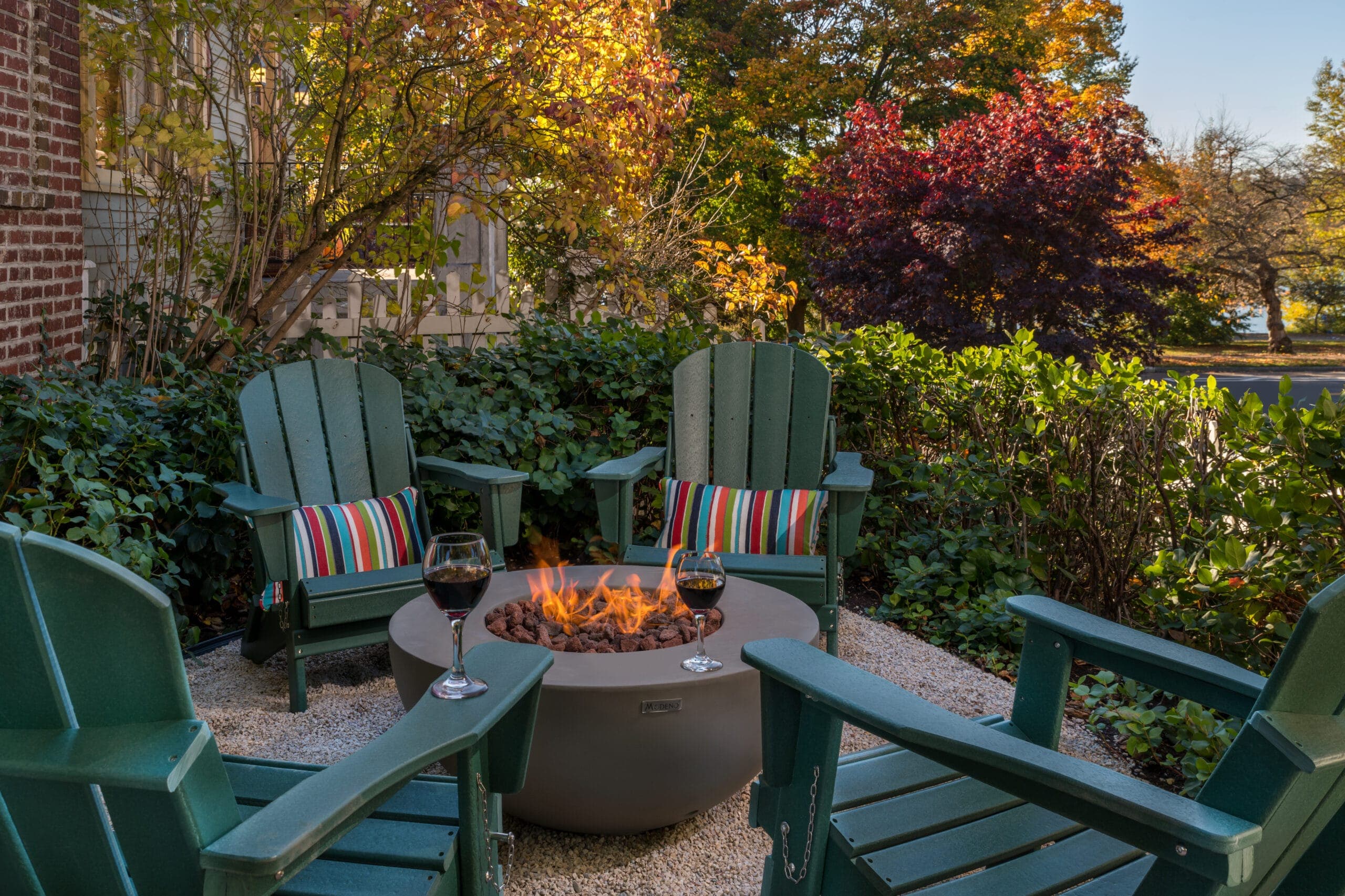 A cozy outdoor seating area with green chairs, a fire pit, and glasses of wine surrounded by colorful autumn foliage.