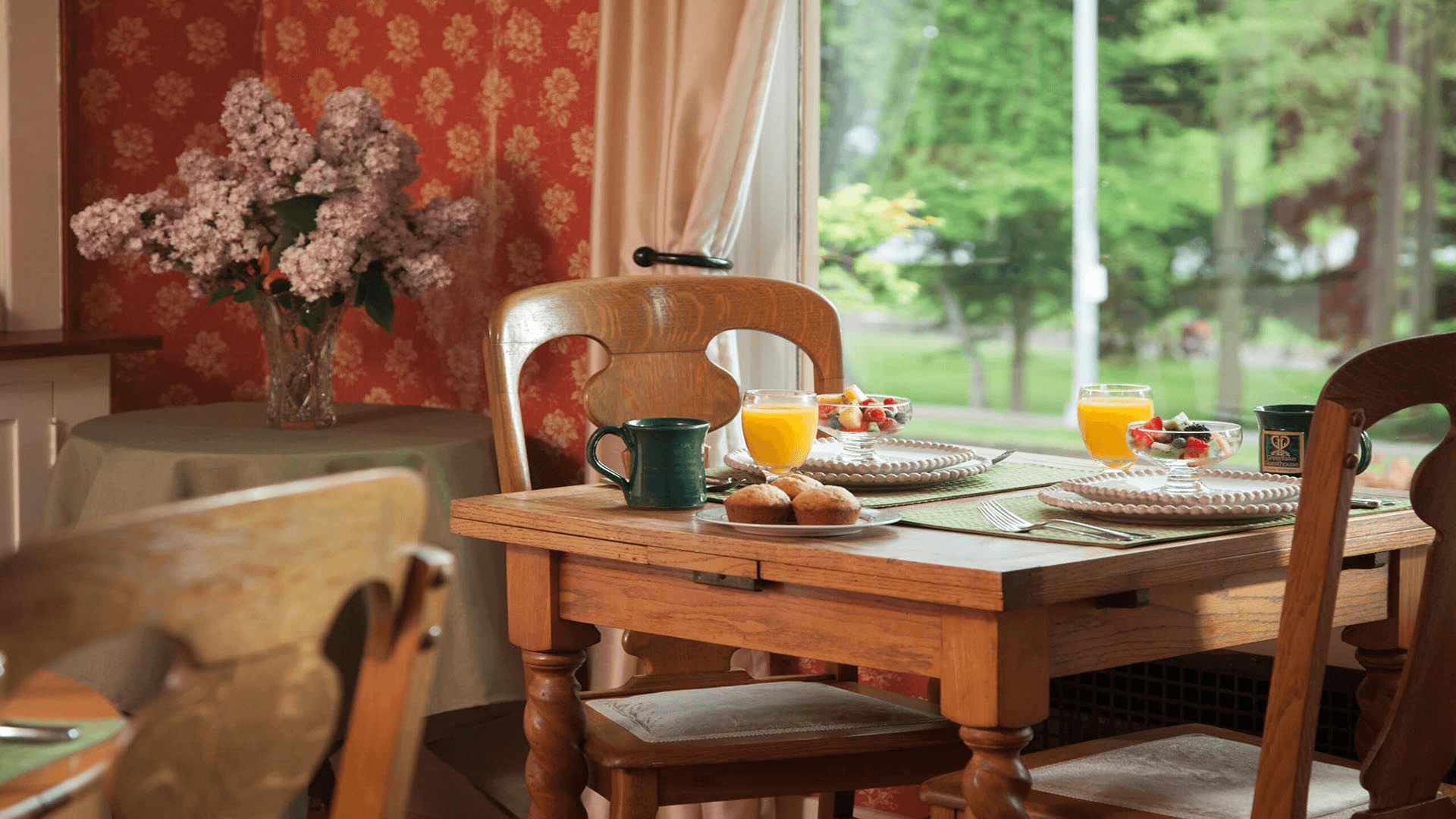 A cozy breakfast table set with fruit, muffins, and beverages near a window with floral decorations.