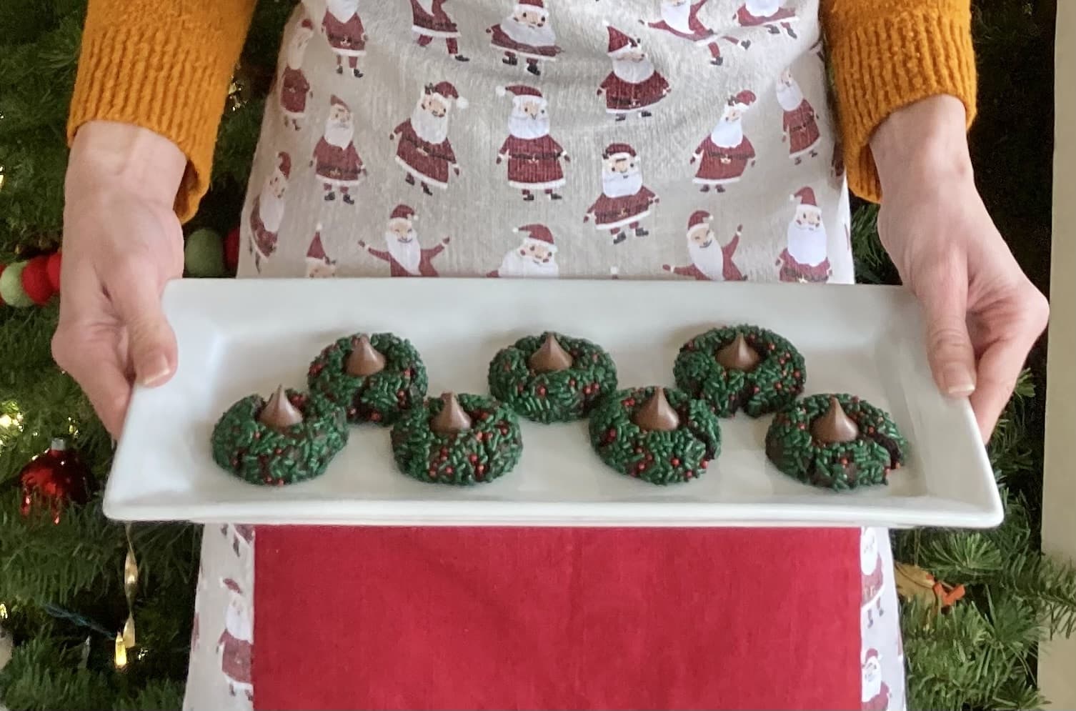 A person holding a platter of festive green wreath cookies adorned with red sprinkles and topped with chocolate kisses.