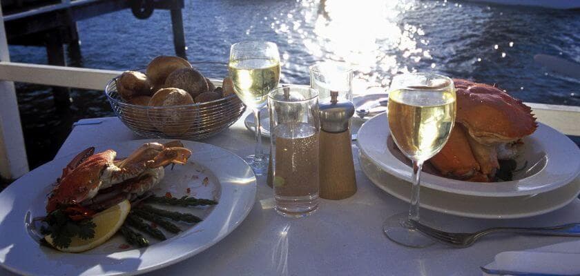 Two plates of crab and asparagus on a table with glasses of white wine, set near a waterfront.