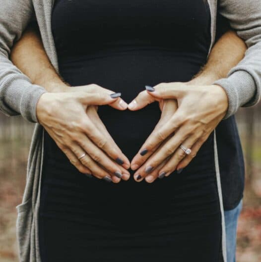 Two hands forming a heart shape over a pregnant belly.