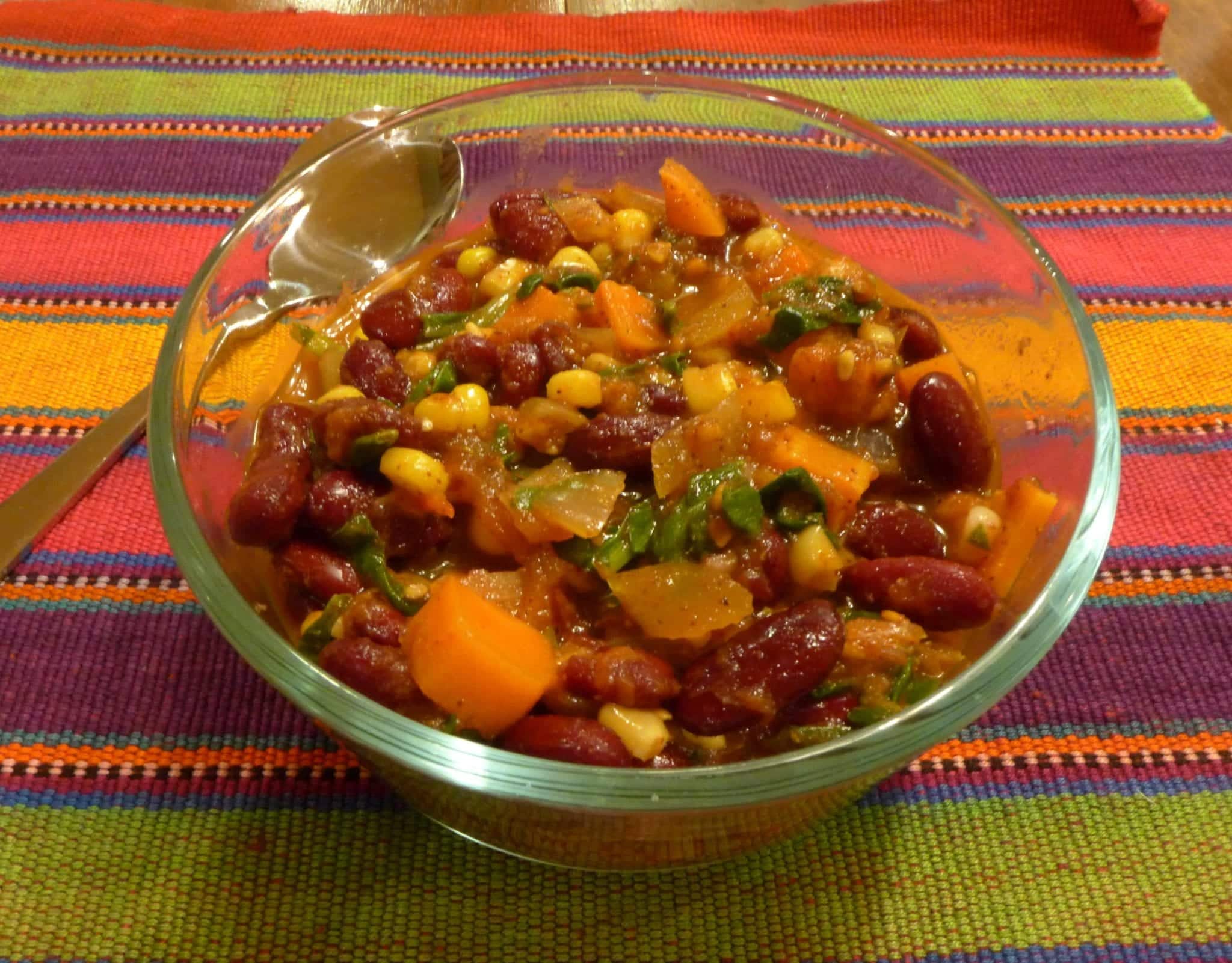 A bowl of colorful bean and vegetable chili sits on a striped cloth with a spoon beside it. A bowl of colorful bean and vegetable chili sits on a striped cloth with a spoon beside it.