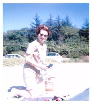A woman with red hair kneels on the beach holding a camera and a handbag, with trees and parked cars in the background.
