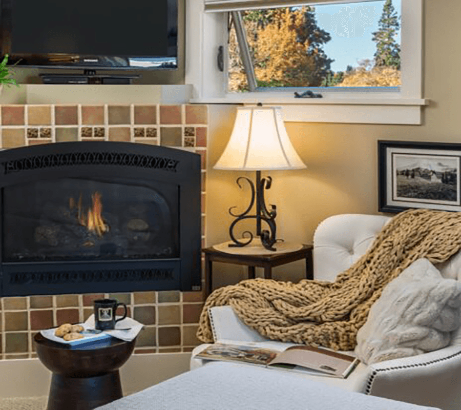 A cozy living room featuring a fireplace, comfortable chair, lamp, and a view of colorful autumn trees outside.