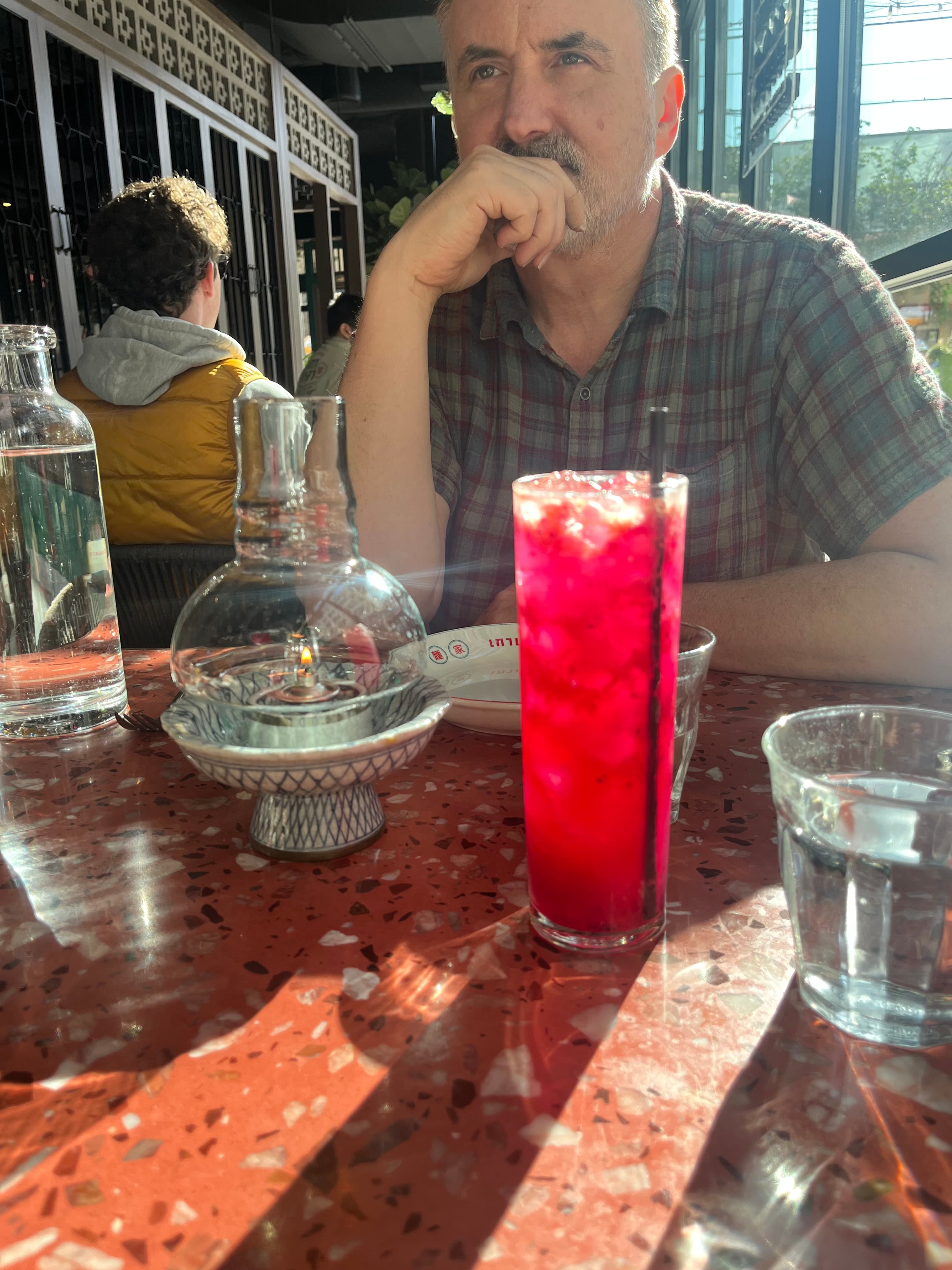 A man with short hair sits at a table, contemplatively resting his chin on his hand, near a vibrant red cocktail and water glasses.