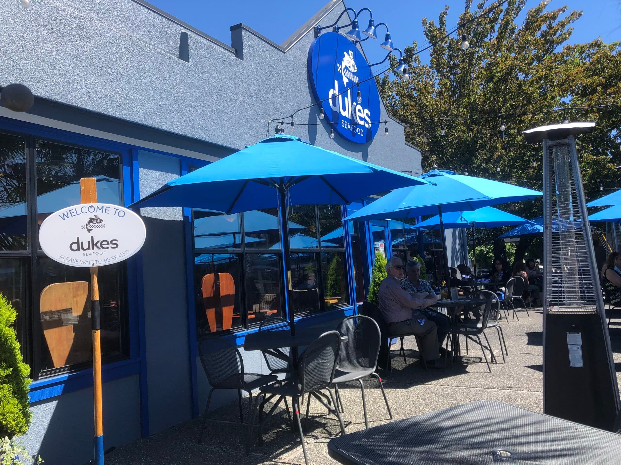 Exterior of Dukes restaurant with blue umbrellas and outdoor seating on a sunny day.