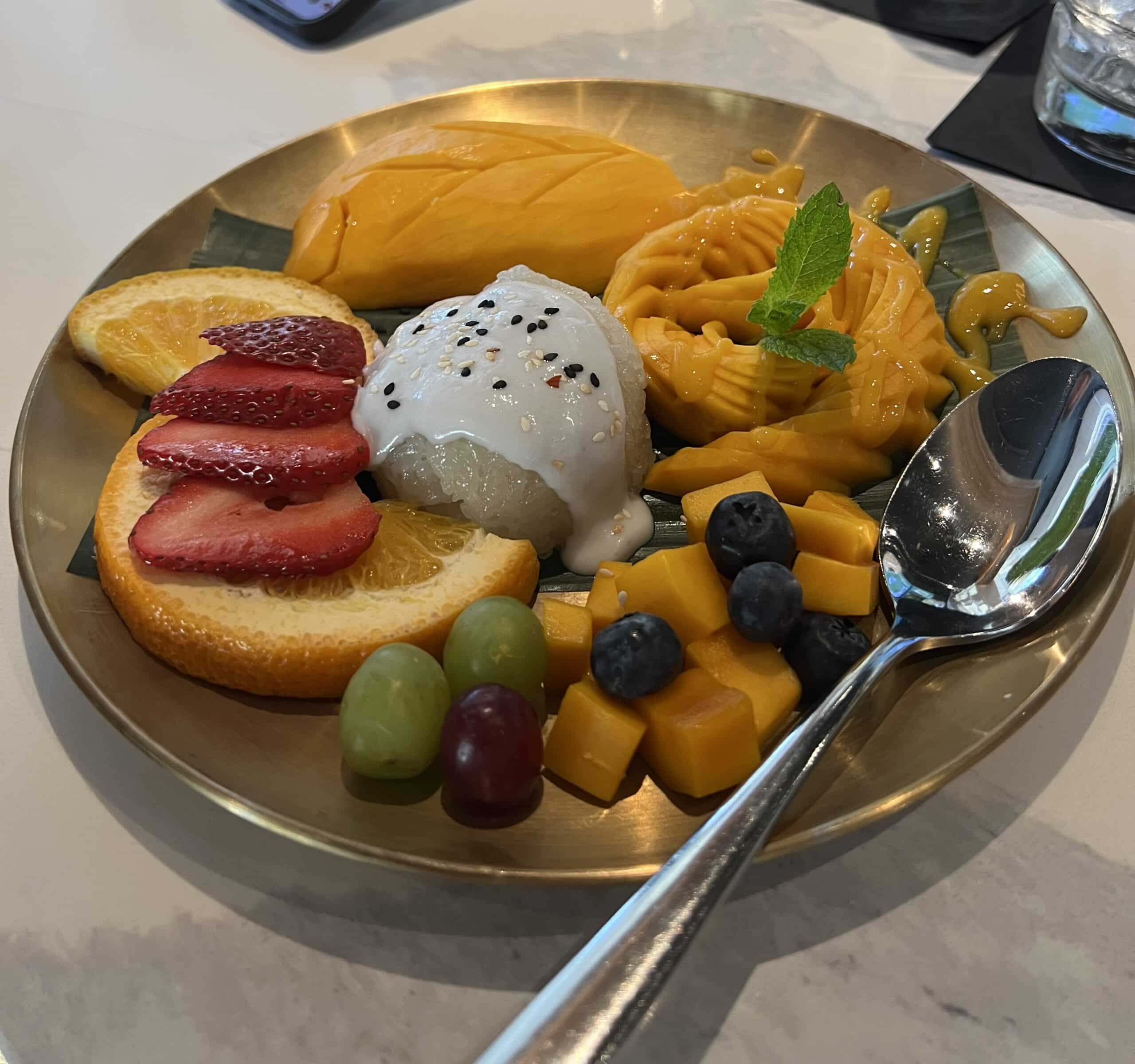 A golden plate filled with fresh fruits, mango slices, a scoop of coconut ice cream, and a sprig of mint.