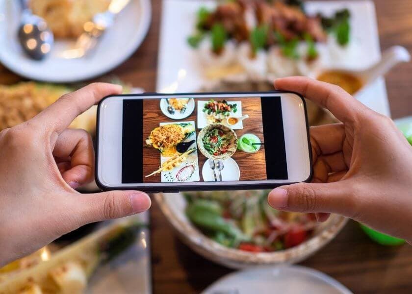A person holds a smartphone displaying photos of food dishes, surrounded by various plates of food on a table. A person holds a smartphone displaying photos of food dishes, surrounded by various plates of food on a table.
