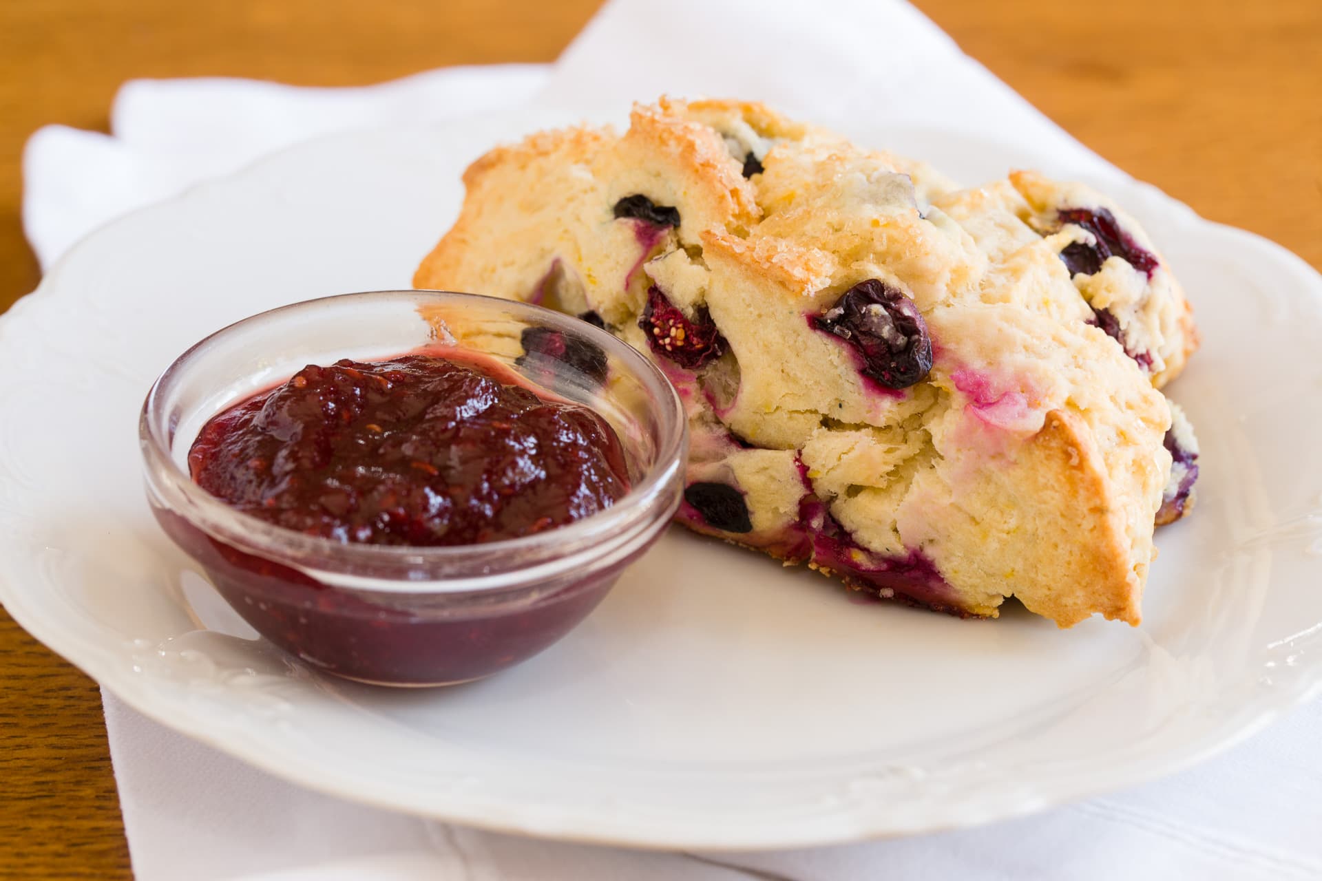 A slice of blueberry scone served with a small bowl of berry jam on a white plate. A slice of blueberry scone served with a small bowl of berry jam on a white plate.