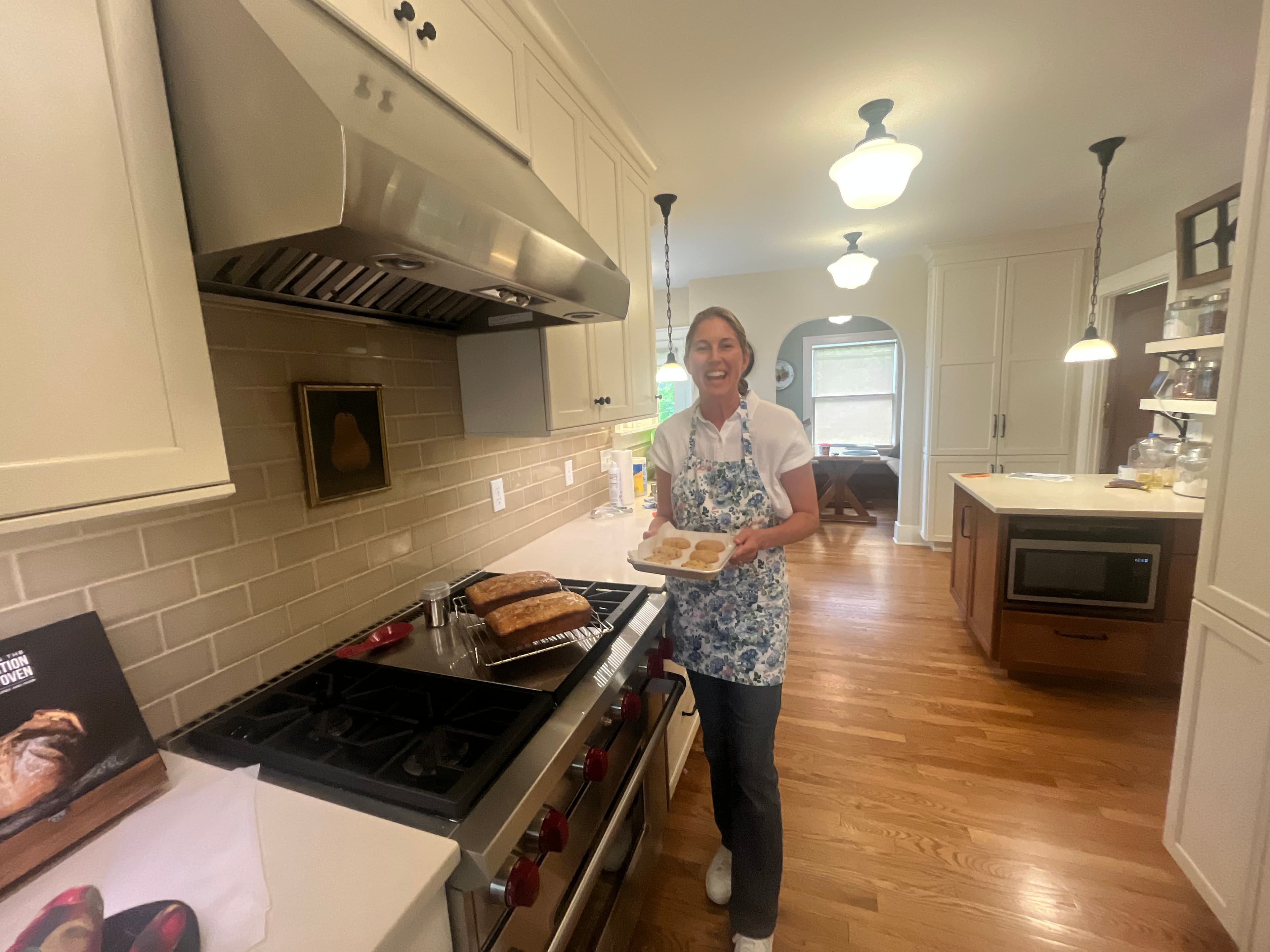 A smiling woman in an apron holds a tray of baked goods in a modern kitchen. A smiling woman in an apron holds a tray of baked goods in a modern kitchen.