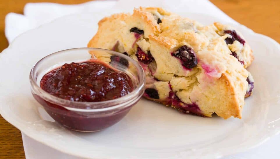 A piece of blueberry scone served with a side of berry jam on a white plate.