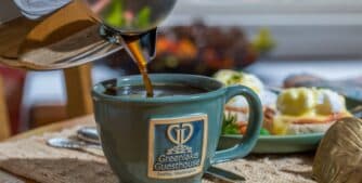 Coffee is being poured into a blue mug with a guesthouse logo, accompanied by a breakfast spread.