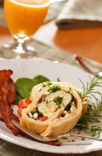 A slice of rolled omelette filled with vegetables and cheese, accompanied by bacon and a glass of orange juice.