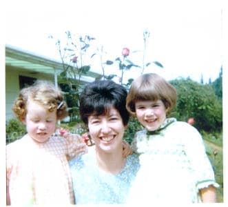 A smiling woman poses with two young girls outdoors among blooming roses.