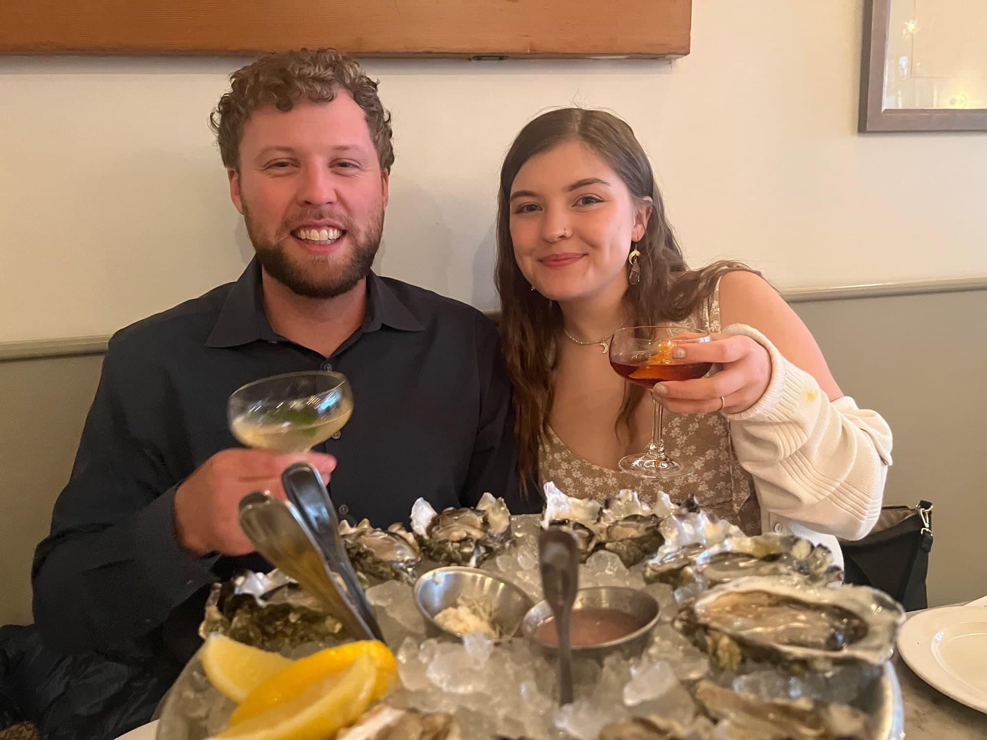 A man and woman smile while holding drinks in front of a platter of oysters at a restaurant.