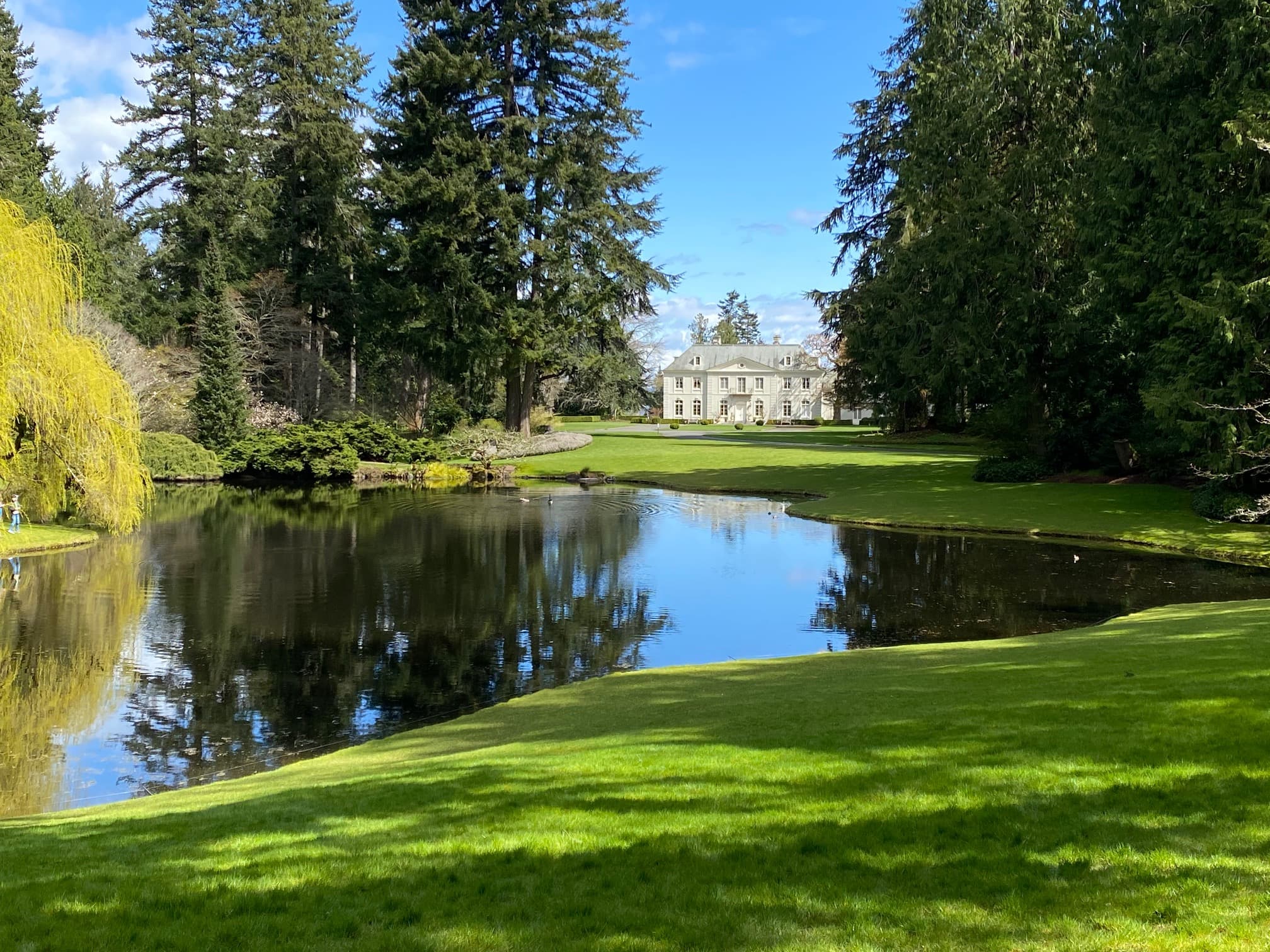 A picturesque mansion reflected in a calm pond, surrounded by lush greenery and tall trees.