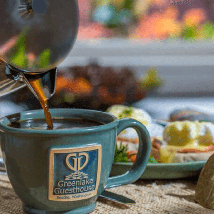 A steaming cup of coffee is being poured from a shiny stovetop pot, alongside a plate of breakfast.