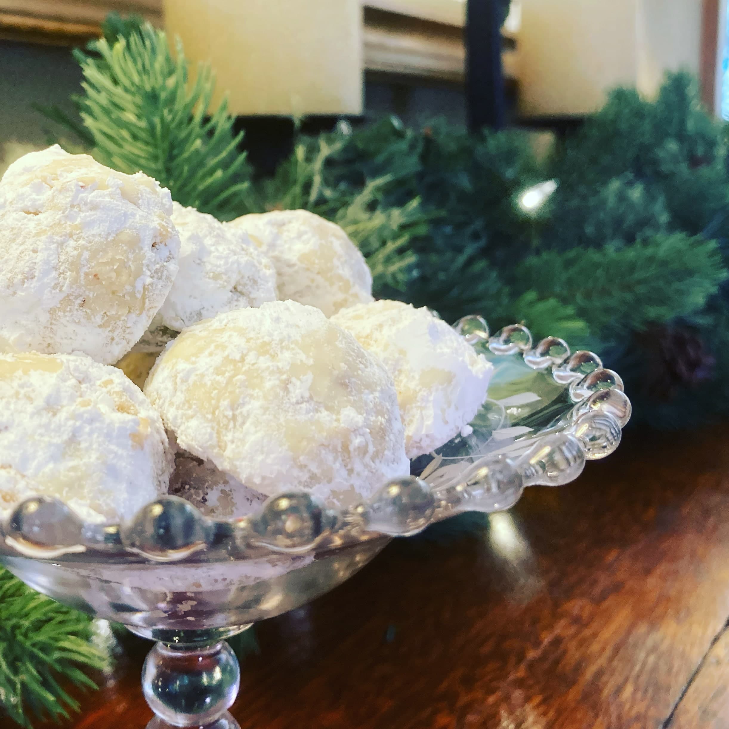 A crystal dish holds powdered sugar-coated cookies, surrounded by evergreen branches.