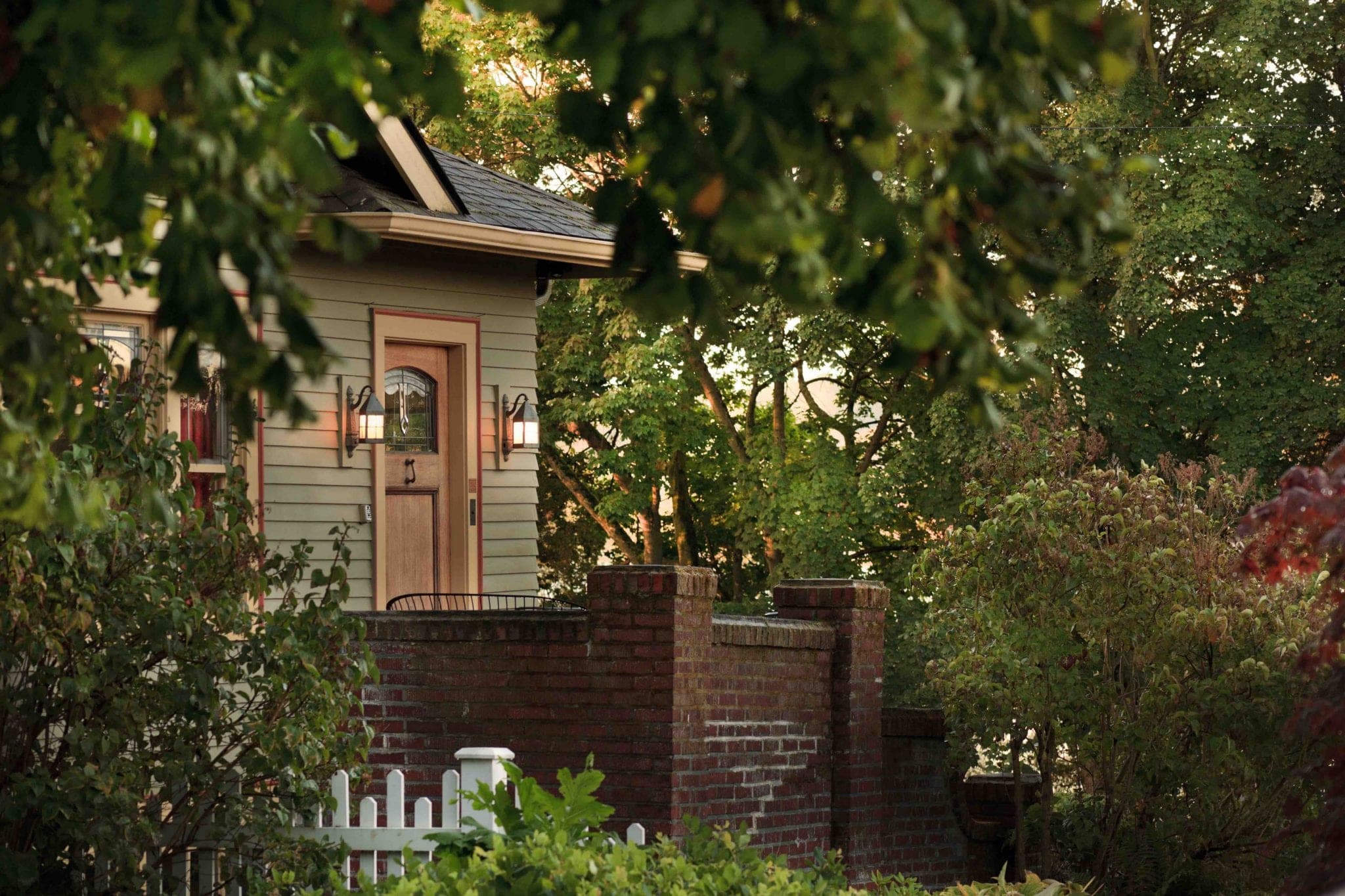 A quaint, green house with a front porch surrounded by lush trees and a brick wall.