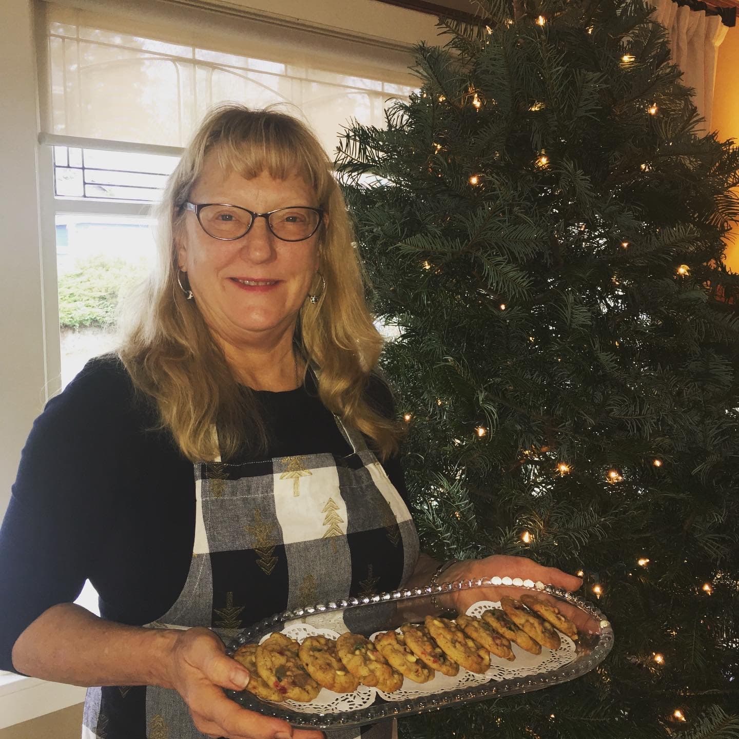A smiling woman holds a tray of cookies in front of a decorated Christmas tree.